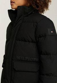 Osoba v černé prošívané bundě s vysokým límcem, klopovými kapsami a logem Tommy Hilfiger na hrudi a rukávu.