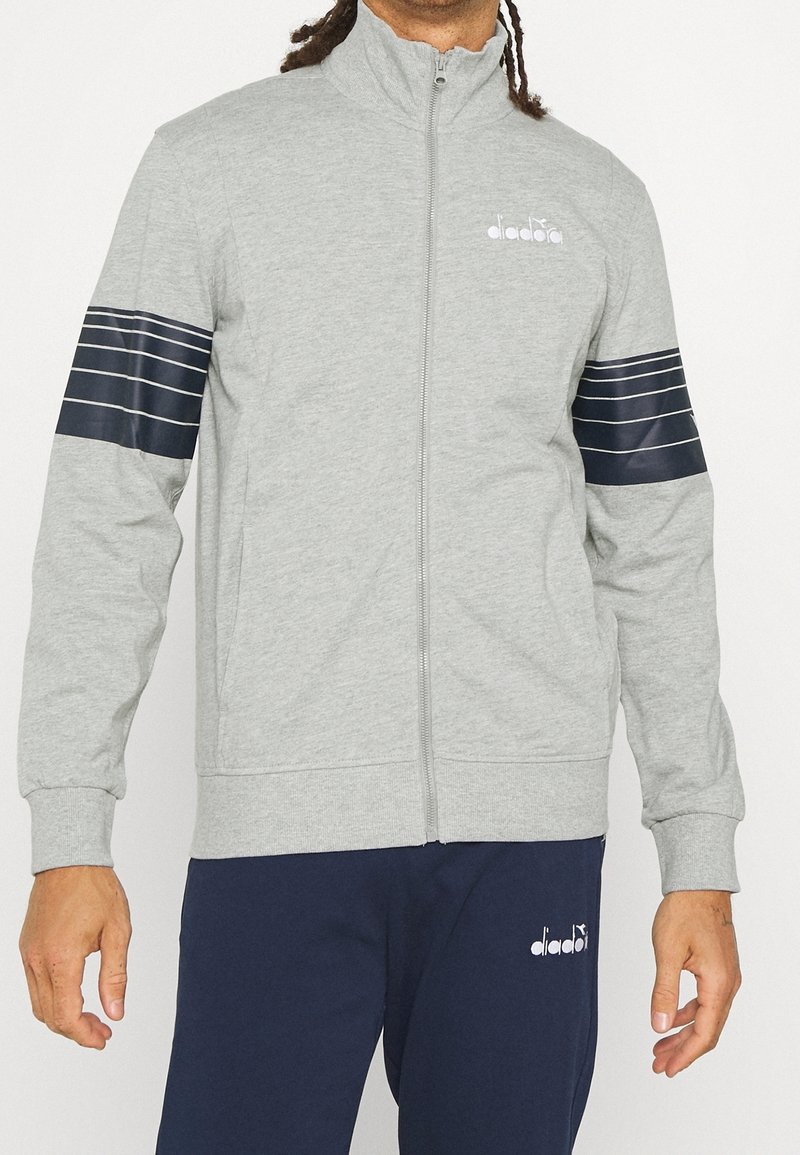 Homme portant une veste zippée gris clair avec des rayures bleu marine sur les manches et un pantalon bleu marine, tous deux avec des logos blancs Diadora.