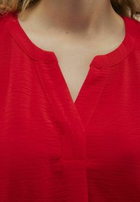 Maglietta rossa con scollatura a V, realizzata in un tessuto strutturato, con cuciture semplici e vestibilità rilassata. Nessun accessorio o motivo particolare.