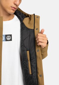 Mann hält braune Jacke offen, um das schwarze Steppfutter zu zeigen, trägt ein weißes Hemd mit zwei Friedenssymbolen in schwarzen und blauen Quadraten.