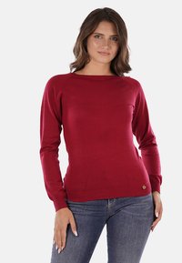 Maglione rosso lavorato a maglia con scollo rotondo e maniche lunghe. Presenta un orlo a coste e un piccolo logo circolare decorativo. Tessuto liscio.