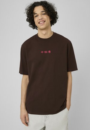 Jeune homme aux cheveux bouclés portant un T-shirt marron avec de petites icônes rouges de feuille, serpent et pot, souriant sur un fond gris uni.