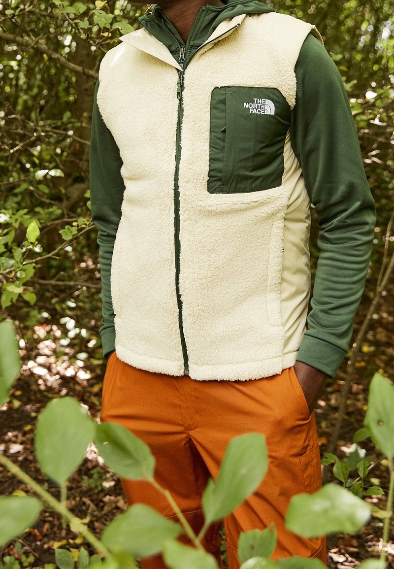Persona che indossa un gilet in pile crema con tasca sul petto verde e logo, camicia a maniche lunghe verde e pantaloni arancioni, in piedi in una foresta verdeggiante.