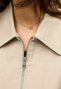 Veste beige zippée avec un col, dotée d'une fermeture éclair argentée. Collier en or avec un pendentif circulaire et un petit détail en perle.