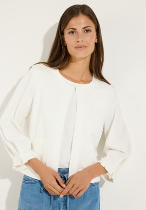 Frau mit langen braunen Haaren, die eine weiße offene Strickjacke über einem weißen Top und blauen Jeans trägt, steht vor einem schlichten Hintergrund.