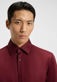 Camicia button-up bordeaux con colletto, texture liscia e bottoni colorati. Presenta un design aderente e nessun motivo o accento visibile.