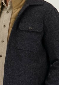 Donkergrijze wollen mix jas met een opgevouwde kraag en een borstzak, gelaagd over een lichtbeige blouse met knopen. Subtiele textuurdetails.