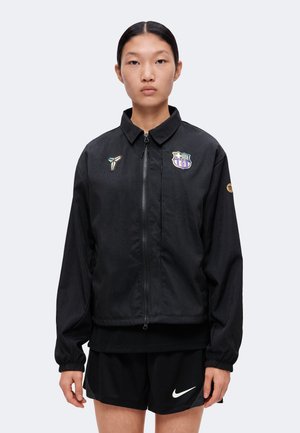Osoba nosiaca čiernu bundu na zips so športovými logami a čierne šortky Nike, stojaca tvárou vpred na jednofarebnom pozadí.