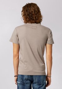 T-shirt taupe a maniche corte con scollo rotondo e un dettagliato quadrato testurizzato sul retro. Abbinato a jeans in denim blu strappati.