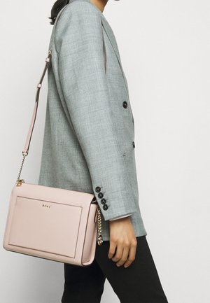Pink leather crossbody bag with a smooth texture, gold chain strap, and front pocket. Worn with a light gray blazer and black pants.