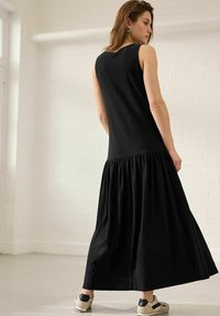 Rochie maxi fără mâneci, de culoare neagră, cu partea inferioară gathered; realizată dintr-un material texturat. Modelul poartă pantofi de nuanță deschisă cu accente negre.