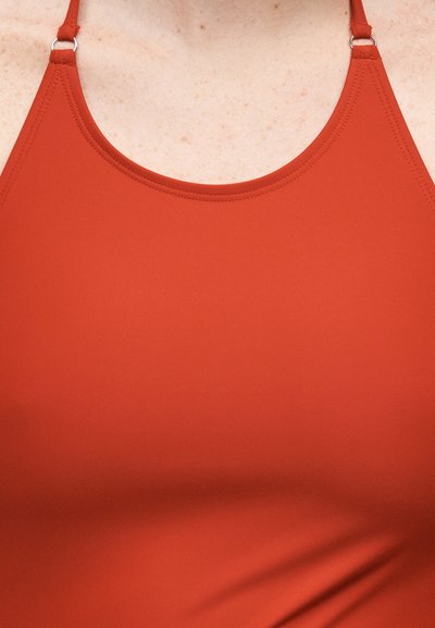 Κοντινό πλάνο ενός ατόμου που φοράει ένα τοπ σκουριάς με λεπτές ρυθμιζόμενες τιράντες και λεπτομερή ραφή κατά μήκος του ντεκολτέ.