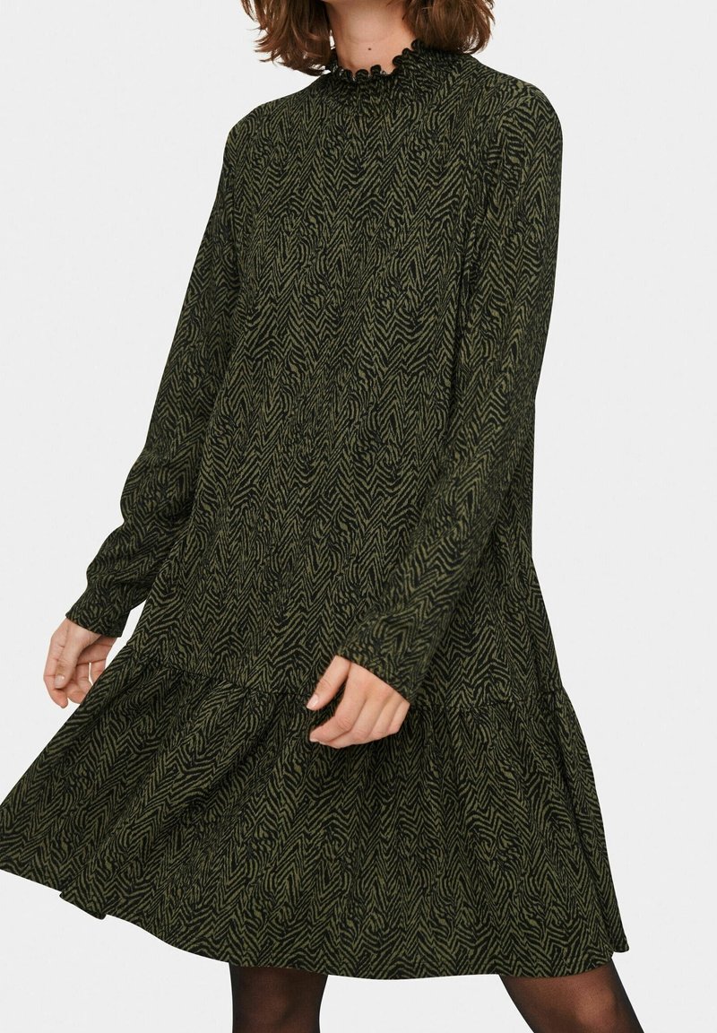 Mörkgrön klänning med långa ärmar i ett tyg med en texturerad zigzag-mönster, som har en volangkrage och en flerskiktad nederkant.