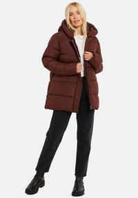 Manteau en duvet marron avec capuche, doté d'une fermeture éclair, d'un design matelassé et de poches latérales. Porté avec un jean noir et de grosses bottes noires.