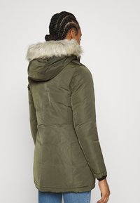 Olivgrön parkas med huva i fuskpäls, med vattentätt yttertyg och lång design. Slät textur och fickor med dragkedja.