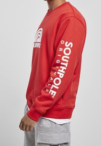 Felpa rossa con logo bianco "SOUTHPOL" e grafica arcobaleno sul davanti, e testo "SOUTHPOL ORIGINALS" lungo la manica sinistra. Materiale in cotone.