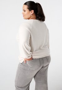 Vrouw met donker haar in een paardenstaart, gekleed in een crèmekleurige trui en grijze jeans, poseert met één hand in haar achterzak.