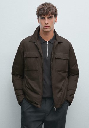 Young man with brown tousled hair wearing a dark brown zip-up jacket over a black polo shirt with a silver zipper.