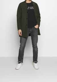 Abrigo ligero verde sobre una camiseta negra con logo, combinado con jeans grises y zapatillas blancas. El abrigo tiene un frente abotonado.