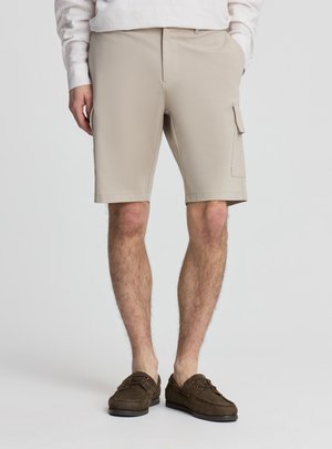 Pantaloncini cargo beige fino al ginocchio indossati con scarpe da barca marroni e camicia chiara a maniche lunghe, mani rilassate lungo i fianchi.