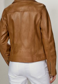 Giacca di pelle marrone con un design strutturato, cuciture in vista e polsini con cerniera. Indossata sopra pantaloni bianchi, che evidenziano una silhouette aderente.