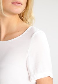 Vit kortärmad topp med rund halsringning. Tillverkad av ett slätt tyg med en subtil lyster. Har upprullade ärmslut och en avslappnad passform.