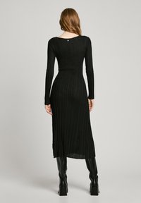 Black ribbed knit dress with long sleeves, fitted silhouette, and a scoop back featuring a small metal accent. Worn with knee-high boots.