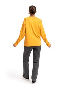 Maglietta gialla a maniche lunghe con collo alto, maniche raglan e cuciture in vista, abbinata a pantaloni grigio scuro e scarpe nere.