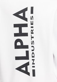 Bela majica s kapuco z velikim črnim napisom "ALPHA INDUSTRIES," ki je postavljen vertikalno po hrbtu. Gladka tekstura blaga.