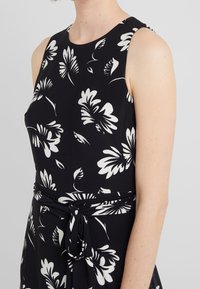 Bezramenné černé šaty s velkým bílým květinovým potiskem, s uvázaným páskem v pase, nošené osobou s světlou pletí.