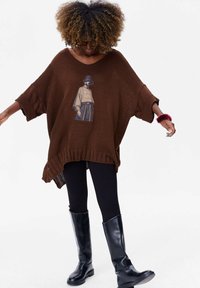 Bruine gebreide poncho met een slouchy silhouet, voorzien van een geïllustreerd ontwerp. Gepaard met zwarte leggings en kniehoge glanzende zwarte laarzen.