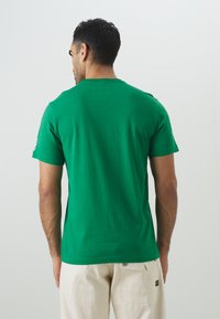 Grøn bomulds t-shirt med rund hals og korte ærmer. Stoffet virker glat med en let stræk, og har et logo på venstre ærme.