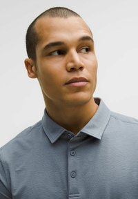 Svetle šedé polo tričko s límečkem, tříknoflíkovou légou a jemnou texturou. Model má krátké vlasy a dívá se mírně na stranu.