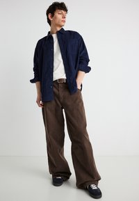 Jeune homme portant une chemise bleu marine à boutons sur un t-shirt blanc, un pantalon large marron et des baskets noires, debout les mains dans les poches.