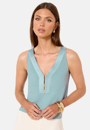 Femeie purtând un top bleu deschis fără mâneci cu fermoar frontal, cercei mici aurii în formă de cerc și două brățări aurii la încheietura mâinii drepte.