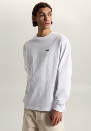 Giovane uomo con capelli scuri fino alle spalle che indossa una maglia bianca a maniche lunghe Vans e pantaloni beige, che guarda di lato.