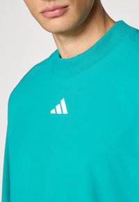 Close-up of person wearing a turquoise sweatshirt with a ribbed collar and a small white Adidas logo on the chest.