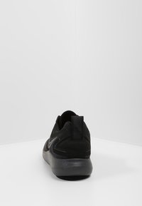 Zapato deportivo negro con un acabado liso y mate; cuenta con una suela gris contrastante, parte trasera texturizada y una lengüeta para facilitar su uso.