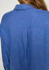 Blå chambray-skjorte med knapkrave, tekstureret stof og en subtil sømdetalje over skuldrene og på ryggen.