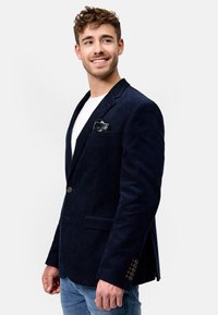 Marineblå cordfløyelsblazer med spisse lapeler, enkel knappelukking og to frontlommer. Har subtile vertikale striper og en lommetørkle.