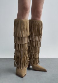 Bottes en suède tan, montantes jusqu'aux genoux, avec des franges superposées, un bout pointu et un talon aiguille. La texture est douce avec une finition mate.
