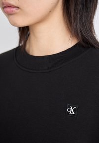 Felpa nera con scollatura rotonda, caratterizzata da un piccolo patch quadrato con il logo "cK" bianco. Il materiale appare morbido con una texture liscia.