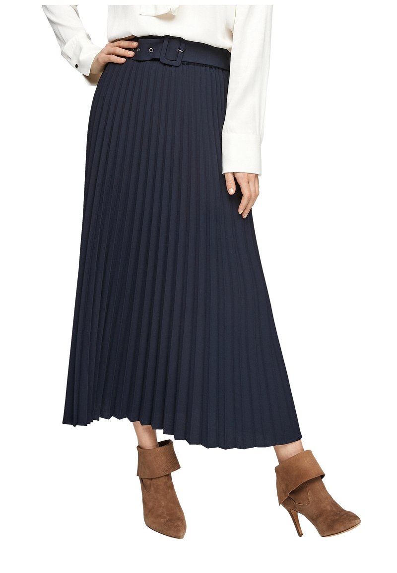 Femme portant une jupe midi plissée bleu marine avec ceinture, un chemisier blanc, et des bottines à talons en daim marron.