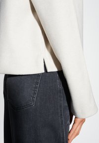 Giacca in pile bianca con vestibilità rilassata, caratterizzata da un orlo dritto e aperture laterali, abbinata a jeans in denim scuro.