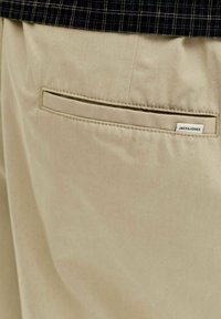 Gros plan sur la poche arrière d'un pantalon beige avec bord cousu et petite étiquette en tissu "JACK&JONES", associée à un tissu à carreaux noir au-dessus.