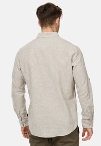 Uomo con camicia beige a maniche lunghe e pantaloni oliva, visto di spalle, su uno sfondo chiaro e uniforme.