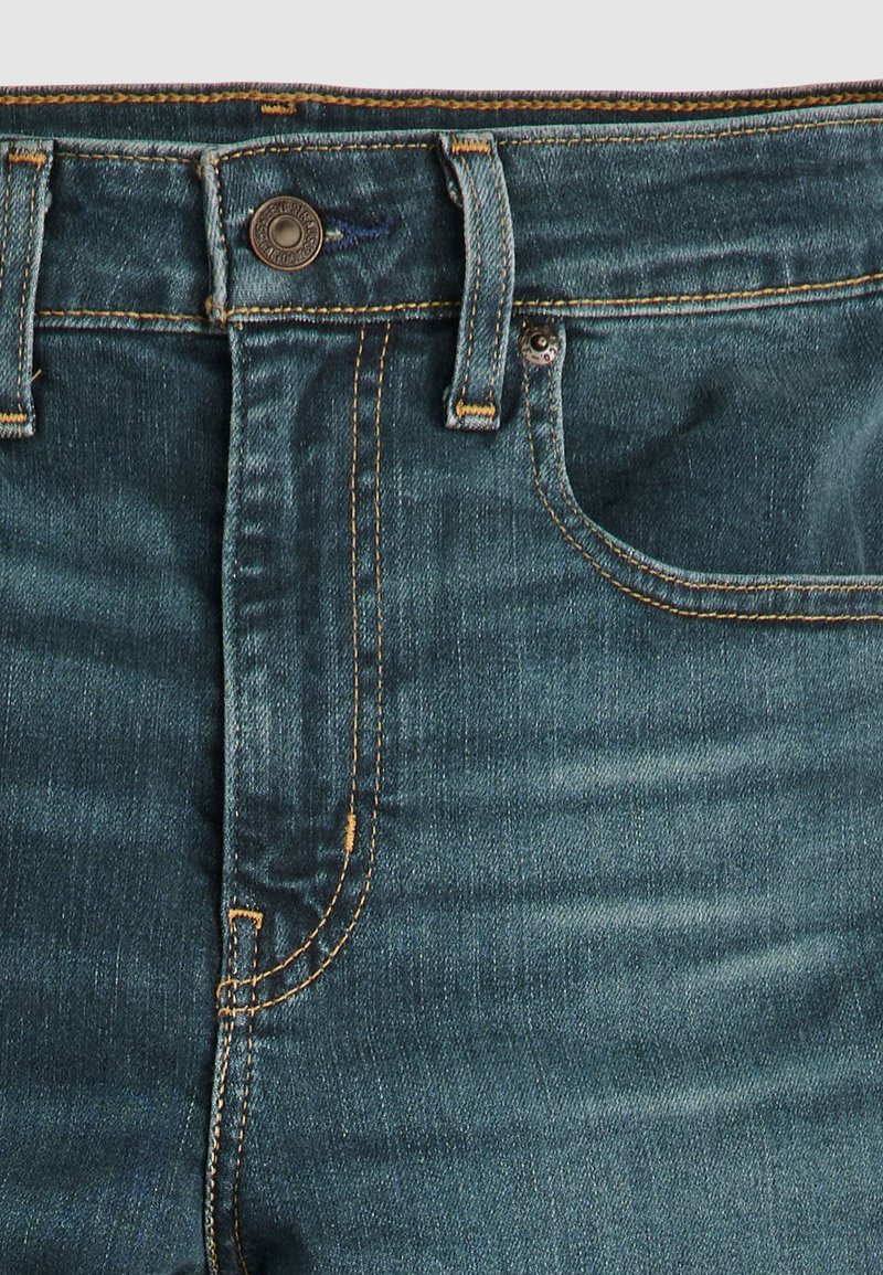Jeans in denim blu scuro con un classico design a cinque tasche, rivetti in rame e cuciture arancioni visibili lungo le cuciture.