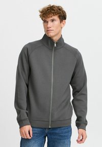 Gray zip-up sweatshirt with a high collar, ribbed cuffs, and a smooth texture, worn over blue jeans. Design features contrasting stitching.