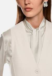 Cremfarbenes ärmelloses Top mit tiefem V-Ausschnitt und einem Knopfverschluss, kombiniert mit einer sanften, hellen Seidenbluse mit gebundenem Kragen.