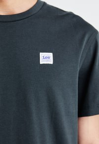 T-shirt grigio scuro a maniche corte con scollo rotondo e un'etichetta quadrata bianca con testo viola che recita "Lee Workwear". Tessuto liscio.